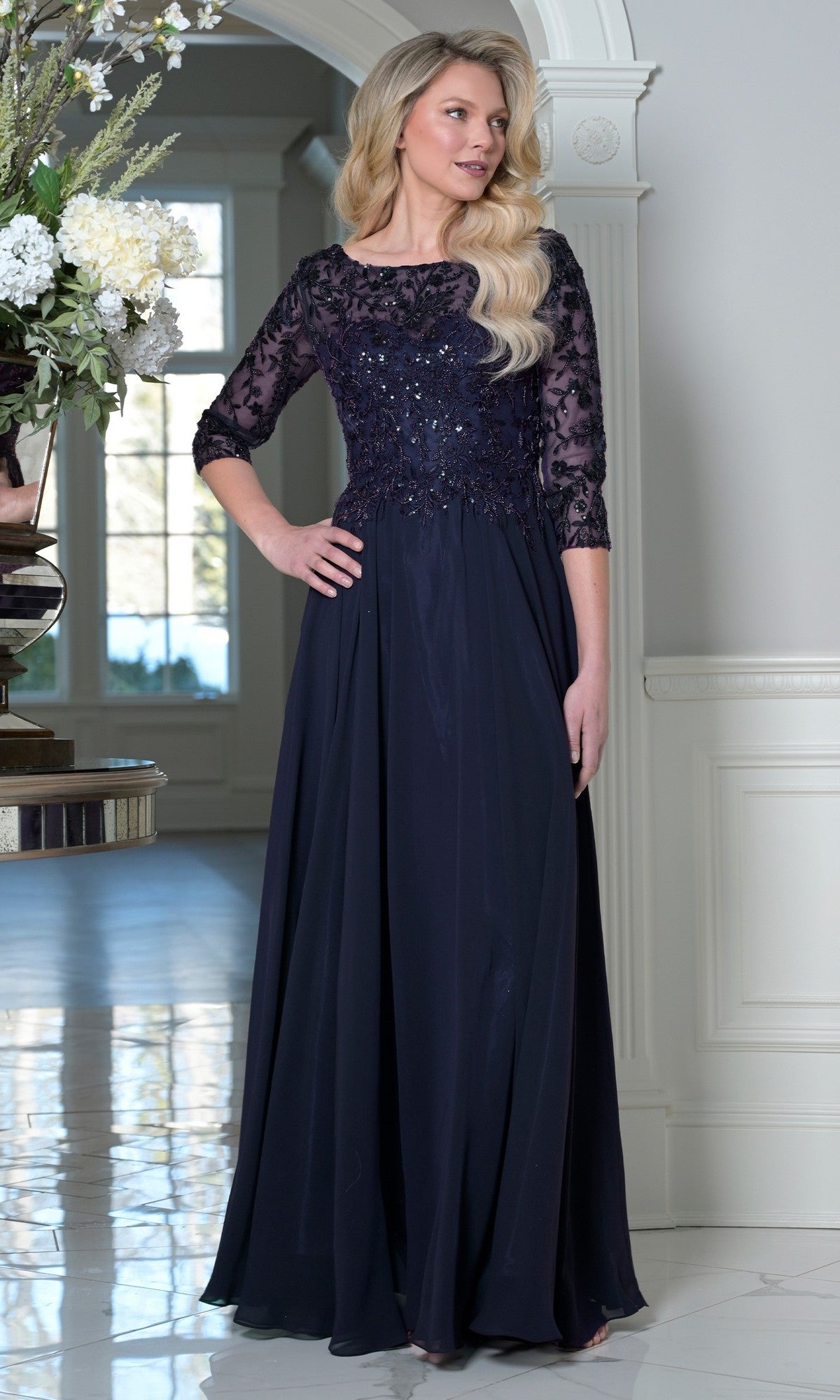 A woman in the navy Marsoni M340SL long formal dress, featuring a beaded-embroidered bodice, lace sleeves, and a flowing A-line chiffon skirt, stands indoors by a large floral arrangement.